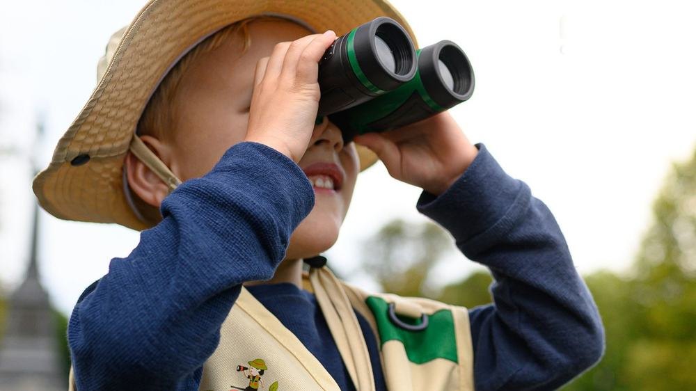 Boy in the explorer vest and hat looking through the binoculars in the park